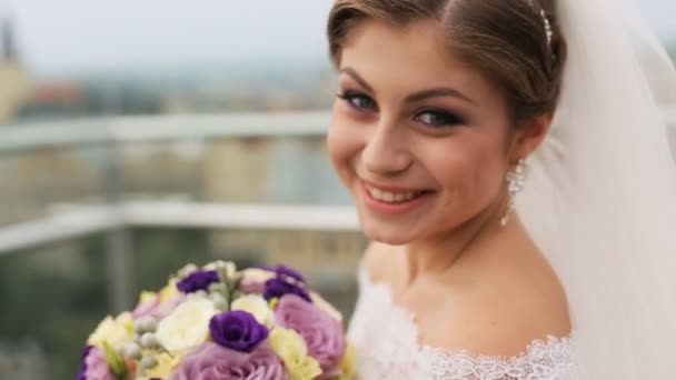 Красивая невеста, пахнущая свадебным цветочным букетом и смотрящая в камеру на размытом фоне старого города. Закрыть — стоковое видео