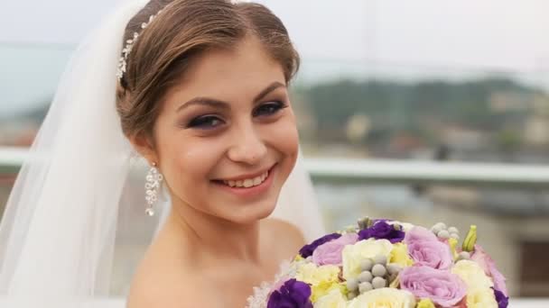 Linda noiva cheirando buquê de flores de casamento e olhando para a câmera no fundo da cidade velha borrada. Fechar — Vídeo de Stock