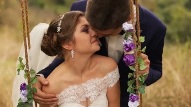 Braut sitzt auf Schaukel in weißem Brautkleid mit Blumenstrauß im Sommerwald, Park. Bräutigam küsst Braut. gerade verheiratet — Stockvideo