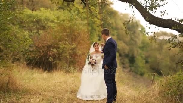 Bruidegom haar mooie bruid is swingend in witte trouwjurk met bloemboeket op schommel in zomer bos, park — Stockvideo