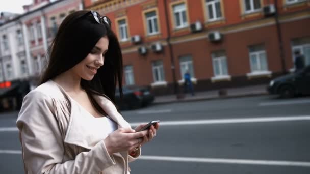 Encantadora joven morena mujer se distrae con mensajes de texto mientras está de pie junto a la calle ocupada — Vídeo de stock