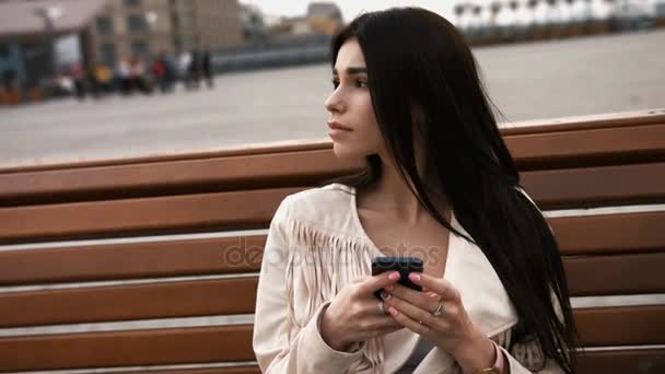 Молодая брюнетка с сотовым телефоном, сидя на скамейке на свежем воздухе, в городской обстановке — стоковое видео