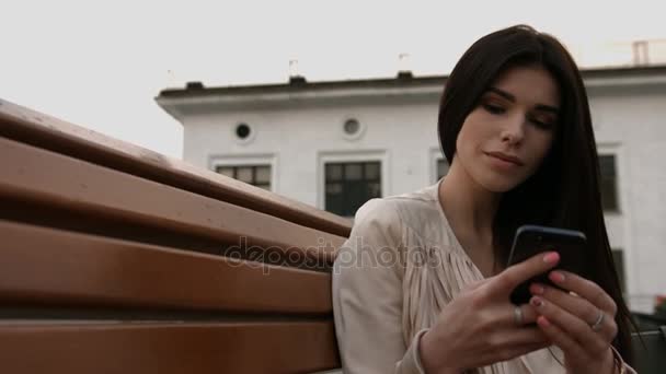 Junge glückliche schöne Frau hält Handy in der Hand und sitzt auf der Bank in der Stadt — Stockvideo