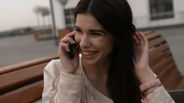 Mujer morena alegre, feliz y sonriente tocando su pelo largo mientras habla en el teléfono celular en la calle, sentada en un banco — Vídeos de Stock