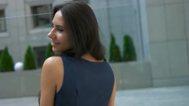 Красивая брюнетка кавказская молодая женщина в синем платье и гуляет по улице, на фоне бизнес-центра. Улыбающаяся девушка . — стоковое видео