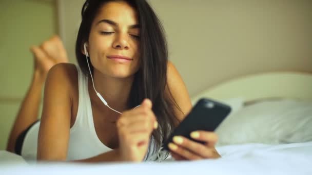 Grappige jongedame liggend op het bed en luisteren naar muziek op haar hoofdtelefoon. — Stockvideo