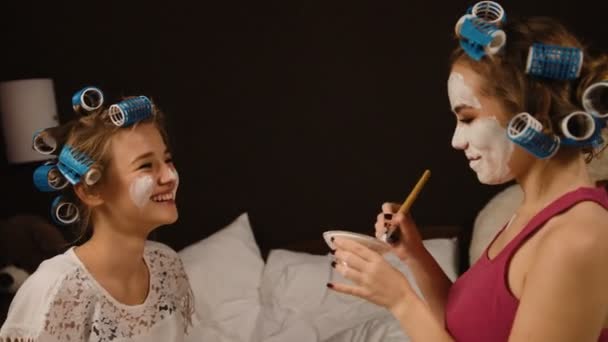 Junge lustige blonde Freundin mit Lockenwicklern macht Maske auf dem Gesicht ihres Freundes. — Stockvideo