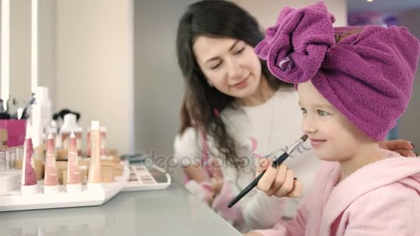 Maquillaje artista aplicar maquillaje con cepillo para niña bonita en el salón blanco. Niña sonriente con bata rosa y toalla en la cabeza — Vídeo de stock