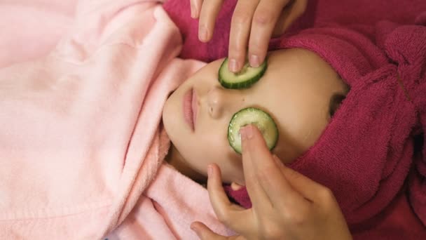 Professional beautician putting cucumber slices on little girls eyes at beauty salon. Spa therapy. Close up view — Stock Video