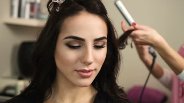 Cabeleireiro profissional, estilista fazendo cachos no cabelo morena das mulheres com ferro de ondulação no salão de beleza . — Vídeo de Stock