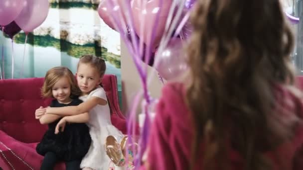 Dos niñas bonitas sentadas en el sofá mientras otra niña trae una caja de regalo con globos. vista posterior — Vídeos de Stock