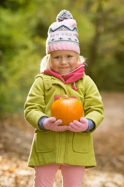 Cute little girl in beanie hat and green coat holding a pumpkin with both hands in the middle of a forest