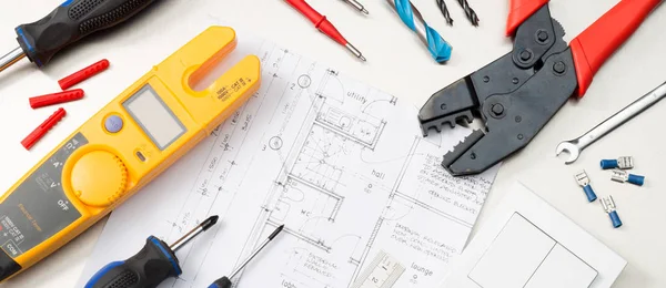Website banner format shot of an assortment of electrical contractors tools on house plans including a multimeter, screwdrivers, wirecutters and a light switch.