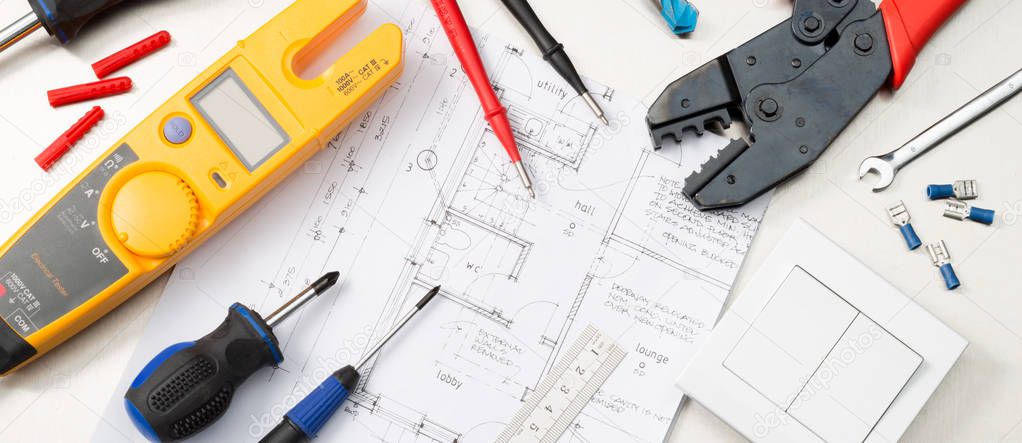 Website banner format shot of an assortment of electrical contractors tools on house plans including a multimeter, screwdrivers, wirecutters and a light switch.