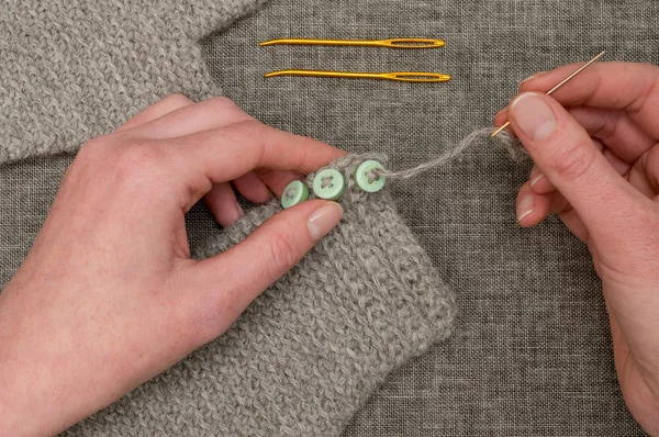 Руки, пришивающие пуговицы к вязанной серой ткани — стоковое фото