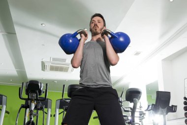 Spor salonunda iki mavi Kettlebells kullanılarak çalışan adam