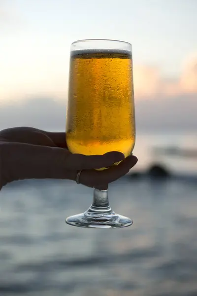 Mano sosteniendo cerveza en copa de vino alto con el océano en el fondo —  Fotos de Stock