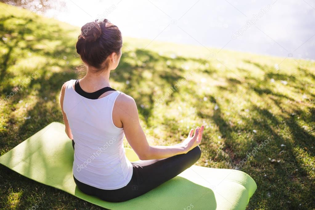 young woman meditating while sitting on yoga carpet in morning park near lake, concept of workout at street  