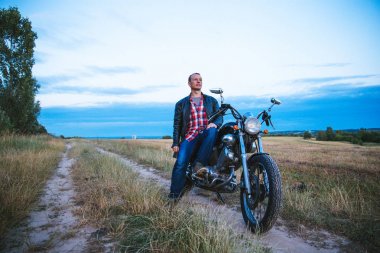 Genç motorcu siyah deri ceket ve kot giyer, onun siyah eski motosiklet üzerinde oturur, beyaz kask tutar ve güzel yumuşak sunset bulvarında görünüyor