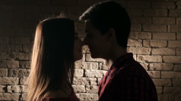Un joven adolescente mira y besa a una chica agradable y feliz con el pelo largo suelto, que toca su mejilla en semidarkness — Vídeos de Stock