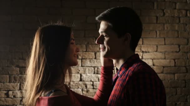 Una buena adolescente con el pelo largo y suelto toca la mejilla de un chico inteligente con corte de pelo de la tripulación y lo besa tiernamente, estar contra una pared de ladrillo — Vídeos de Stock