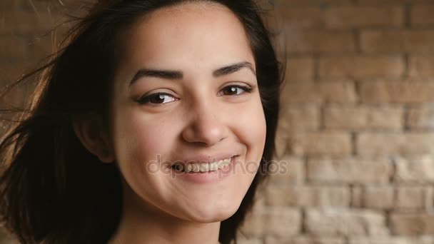 Een geweldig 18-jarige meisje in een rode jurk met open schouders glimlacht raadselachtig en deelt haar positieve emoties terwijl ze in de studio, tegen een bakstenen muur, in slo-mo — Stockvideo
