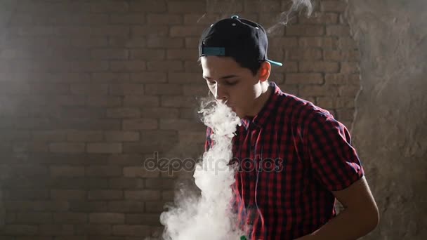 Un adolescente con gorra de béisbol y camisa a cuadros fuma un cigarrillo electrónico haciendo círculos de humo y envía uno grande con la mano a una cámara, mientras está contra una pared de ladrillo . — Vídeo de stock