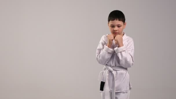 Um pequeno karateka treina golpes de mão, vestido com um quimono branco e um cinto branco, em estúdio — Vídeo de Stock