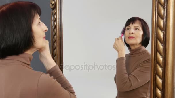 Модная пожилая женщина расчесывает свои красивые каштановые волосы перед художественным зеркалом — стоковое видео