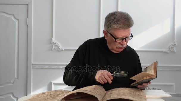 Un hombre mayor explora un manuscrito antiguo y usa un libro viejo y una lente de mano — Vídeo de stock