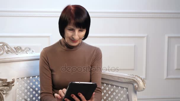 Una mujer de cabeza marrón con una camisa de cuello alto navega por la red en su tableta y sonríe — Vídeo de stock