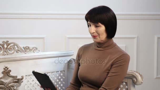 Eine nette Seniorin sitzt im Dreivierteltakt und surft mit ihrem Tablet im Netz — Stockvideo