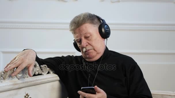 Un hombre mayor escucha la música, está en auriculares, y mira su móvil — Vídeo de stock