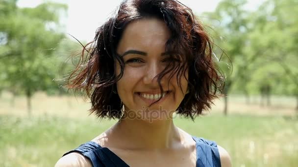 Young woman smiles sincerely on a lawn with green trees on a sunny day in slo-mo — Stock Video