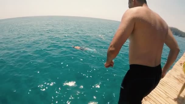 Man springt in het sprankelende zeewater vanaf een pier en verdwijnt er in slo-mo — Stockvideo