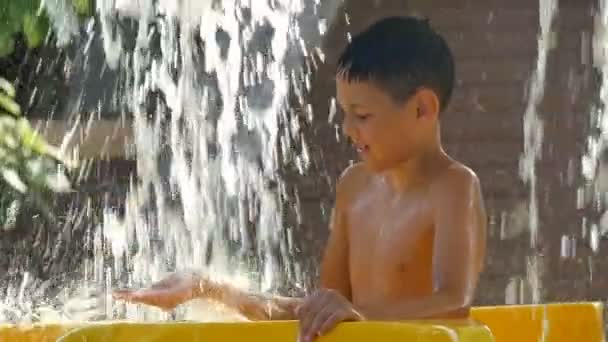 Menino feliz em um dia quente de verão fica sob os riachos de água no parque aquático — Vídeo de Stock