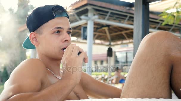 Jonge man rookt een e-sigaret zittend in een zee-bar in de zomer in slo-mo — Stockvideo