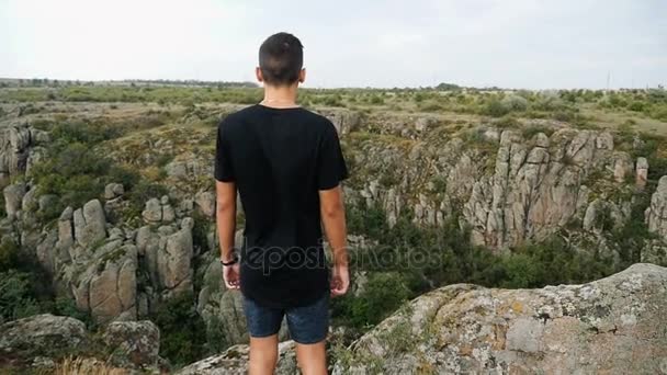 A fiatalember egy sziklán áll, úgy néz ki, egy mély szakadék a hegyekkel a közelben slo-Mo — Stock videók