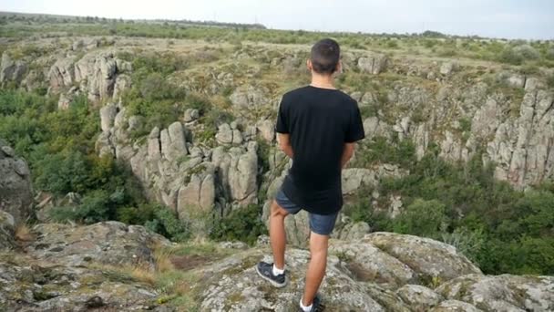 Een jonge man staat bovenop van een hoge klif op zoek op schilderachtige heuvels in slo-mo — Stockvideo