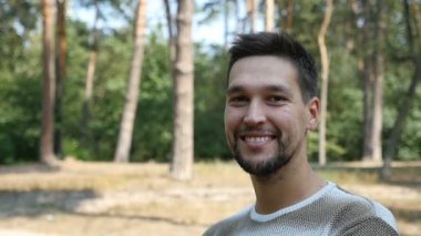 Ağır çekimde yaz aylarında bir çam ormanı içinde güneşli bir çimenlikte genç bir adam gülümsüyor