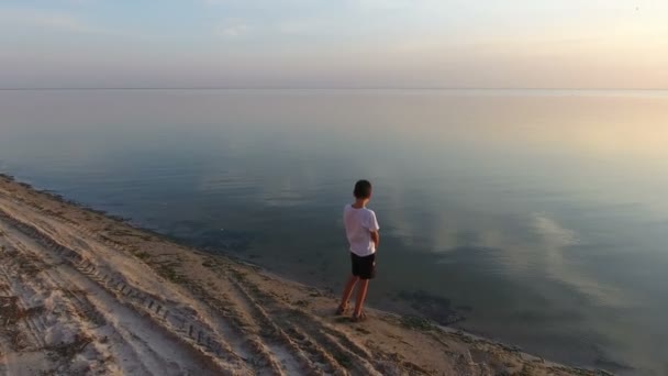 Small boy stands on a rough seashore and enjoys the seascape shot from a drone — Stock Video