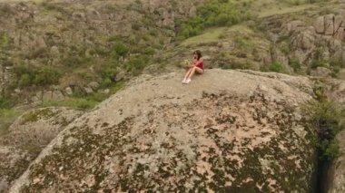 Genç bir kız iyimser bir bakış kırmızı bir tshirt ve güneşli bir günde split kayalar ile kaplı bir dağ tepe üzerinde oturan şort giymiş.  