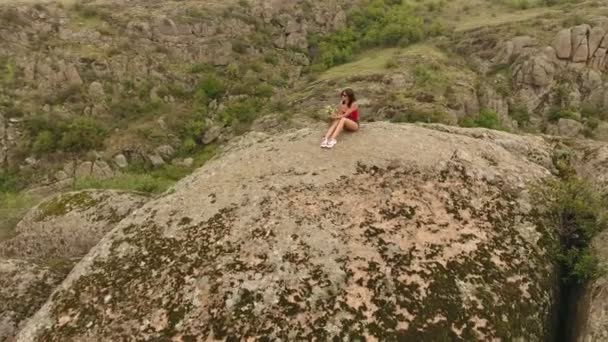 シャツと晴れた日に分割岩で覆われている山のピークの上に座ってパンツに身を包んだ少女の楽観的な見方 — ストック動画