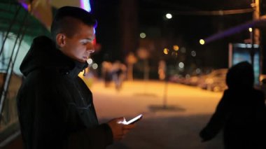 Genç adam anlamına gelir ve mobil ekran dokunaklı ve bir semt akşamları cadde üzerinde bir bilgi arayan kulaklıkları ile bir şehir Kış A genç bir adam profilinde sokak smartphone ekranda bilgi istiyor. Xmas hediye hakkında düşünüyor.