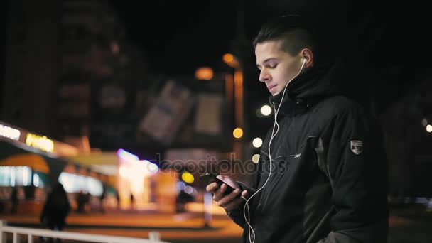 若い男は Earbudstouching 携帯電話や冬の夜の街にいくつかの情報を求めての画面を持つ若い男の冬 見事なクローズ アップの街でスマート フォンの画面に情報を求めています — ストック動画