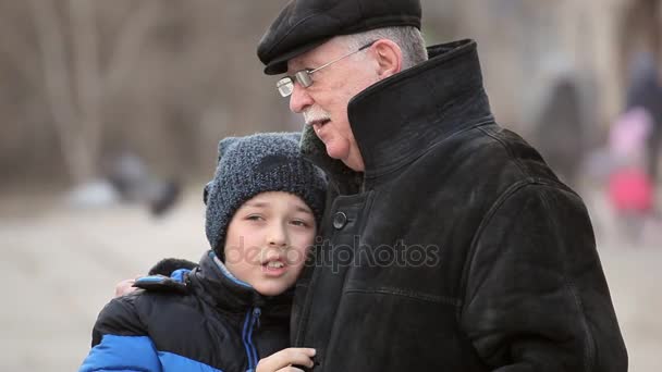 Malý chlapec v legračním klobouku mluví k jeho moudrý dědeček v park lane v zimě potěšující pohled devítileté dítě v vlněná čepice s černou koulí a jeho moudré šedovlasé grand táta, který obejme ho v park lane