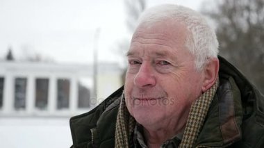 Gri başlı ihtiyar kameraman ve gülümsüyor biraz kışın standları bir koyu yeşil anorak giyinmiş bir gri saçlı adam görkemli bir görünüm üzerinde bir şehir sokak arabalar kışın hareketli döner. O döner ve gülümsüyor.  