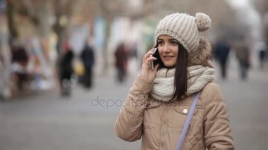 Güzel kız gülümsüyor ve gösterişli bir ara sokakta ona akıllı telefon üzerinde kışın gülümsüyor ve konuşuyor örme şapka neşeli topları ile içinde güzel bir kız şaşırtıcı bir görünümü üzerinde akıllı telefonunu bir sanatsal sokak kışın konuşuyor 