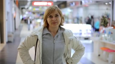 Neşeli sarışın kadın anlamına gelir ve yukarı jestleri anlamına gelir ve yukarı jestleri bir alışveriş merkezinde gösterir gülümseyen bir sarışın kadınla bir alışveriş merkezi A neşeli görünümünü gösterir. O bir beyaz ceket ve gri zipup gömlek..