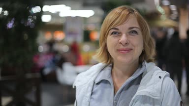 Mutlu sarışın kadın ayakları ve bir alışveriş merkezi ile insanlar duran bir gülümseyen neşeli görüş-in sarışın kadın gülümsüyor ve gülümsüyor yerel alışveriş merkezi. O bir beyaz ceket ve Tshirt kadar gri ZIP mevcuttur.